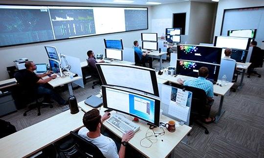 Seven men sitting at desks with large computer monitors analyzing data